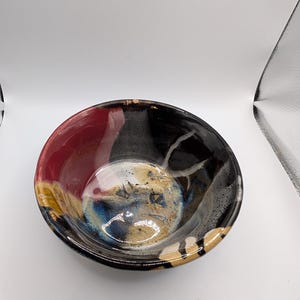 Może przedstawiać: Okrągła ceramiczna miska z błyszczącym wykończeniem, łącząca głęboką czerń, żywą czerwień i odcienie ziemi. Wnętrze zdobi wirujący, marmurkowy wzór z smugami niebieskiego i białego szkliwa, tworząc wielobarwną powierzchnię na całej misce.