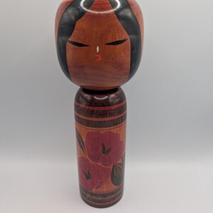 Op de afbeelding: Een houten Kokeshi-pop met een groot, rond hoofd met zwart geschilderd haar, gesloten ogen en een kleine rode mond. Het cilindrische lichaam is versierd met donkerrode bloemmotieven en horizontale zwarte lijnen op natuurlijk hout.