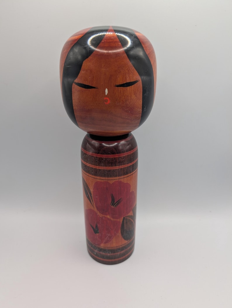 Op de afbeelding: Een houten Kokeshi-pop met een groot, rond hoofd met zwart geschilderd haar, gesloten ogen en een kleine rode mond. Het cilindrische lichaam is versierd met donkerrode bloemmotieven en horizontale zwarte lijnen op natuurlijk hout.