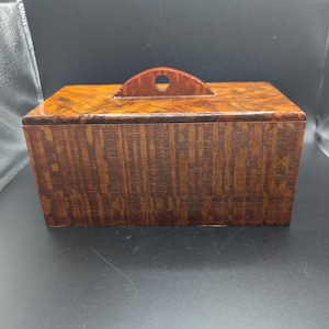 May include: A rectangular wooden box with a dark brown finish and a distinct grain pattern. The lid features a semi-circular handle with a small circular cutout in the center. The box has a smooth, polished surface and a structured, boxy shape.