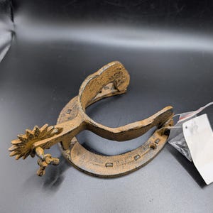 May include: A cast iron boot spur featuring a textured, dark bronze-colored finish. The object consists of a U-shaped heel band integrated with a rowel, which is a small, star-shaped wheel with multiple points, attached to the end of the shank.