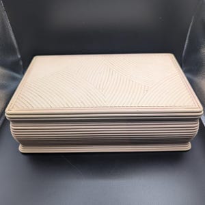 May include: A rectangular, light beige storage box with a lid featuring a textured, repeating curved line pattern. The sides of the box are constructed with multiple horizontal, stacked layers, creating a ribbed appearance around the entire perimeter.