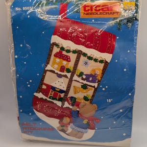 May include: A Bucilla Needlecraft felt stocking kit in a clear plastic package. The kit features a 15 inch stocking design with a red roof, a house, a doll, a sailboat, and a teddy bear. The blue background has white dots and the text No. 85033.