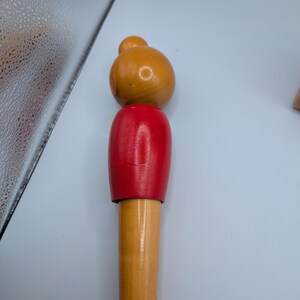 Op de afbeelding: Een houten speelfiguur met een afgeronde kop en een kleinere bol erbovenop, geschilderd in tinten oranje en rood. De figuur heeft een cilindrisch lichaam en staat op een kleine, vierkante houten basis. Het speelgoed is gemaakt van hout en heeft een gladde afwerking.