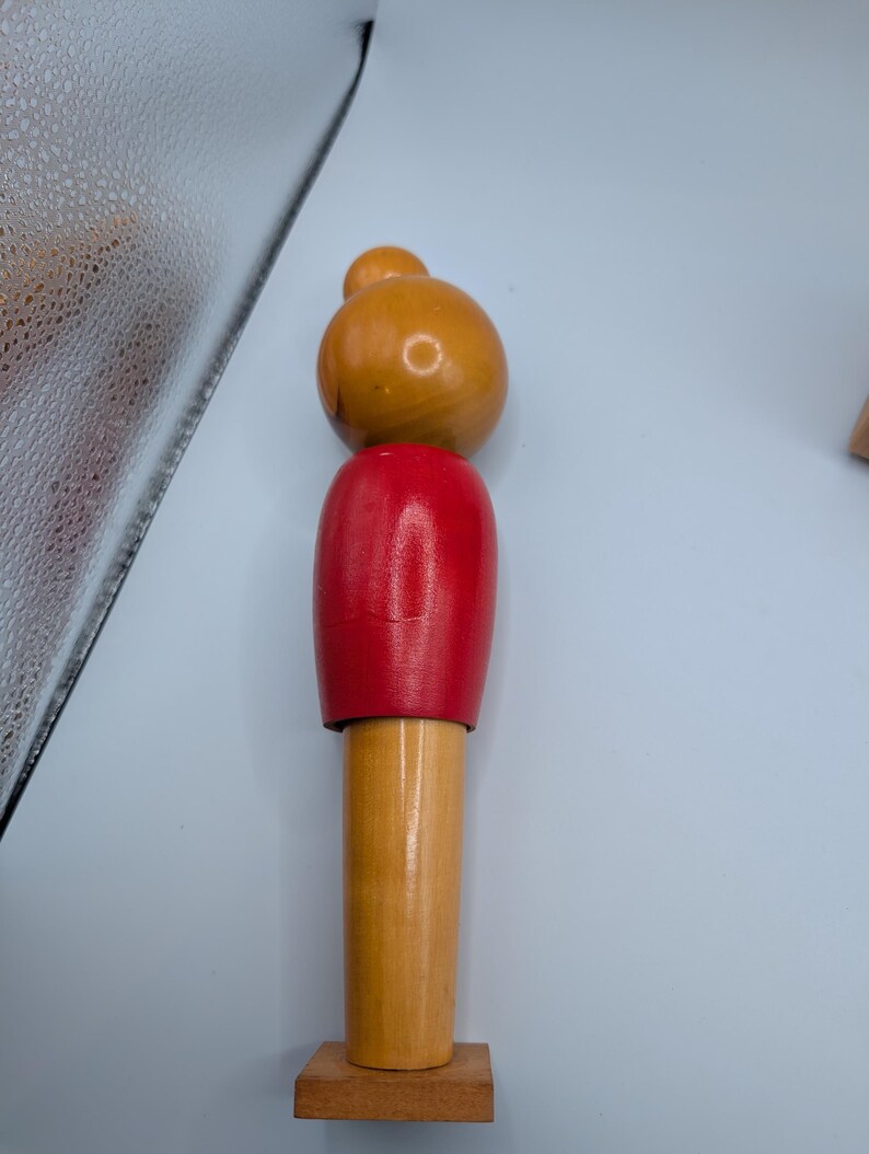 Op de afbeelding: Een houten speelfiguur met een afgeronde kop en een kleinere bol erbovenop, geschilderd in tinten oranje en rood. De figuur heeft een cilindrisch lichaam en staat op een kleine, vierkante houten basis. Het speelgoed is gemaakt van hout en heeft een gladde afwerking.