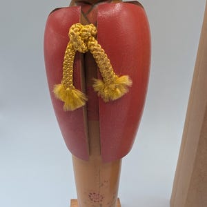 Op de afbeelding: Houten Japanse Kokeshi-pop met een rode kimono-achtige top en een gele koord. De pop heeft een lichtbruin lichaam met bloemmotieven en is gemonteerd op een houten voet. De kop is donkerbruin.