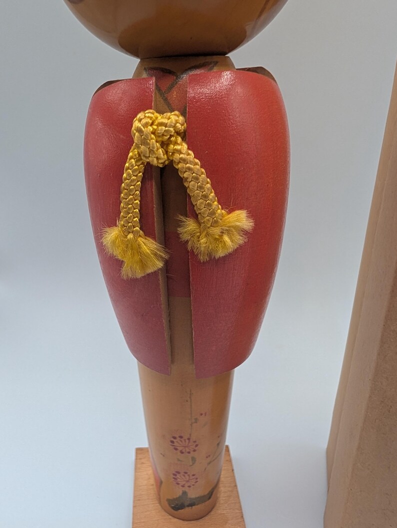 Op de afbeelding: Houten Japanse Kokeshi-pop met een rode kimono-achtige top en een gele koord. De pop heeft een lichtbruin lichaam met bloemmotieven en is gemonteerd op een houten voet. De kop is donkerbruin.