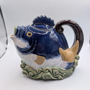 Peut inclure: Une théière en céramique en forme de poisson avec un corps bleu foncé brillant, un ventre blanc et une anse marron. Le poisson présente un couvercle en forme de nageoire dorsale, une queue beige et repose sur une base sculptée en vagues vertes.