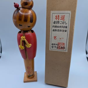 Op de afbeelding: Een houten Japanse Kokeshi-pop met een rode jas en een bruin hoofd. De pop staat naast een bruine doos met Japanse tekst. De pop is ongeveer 15 cm hoog.