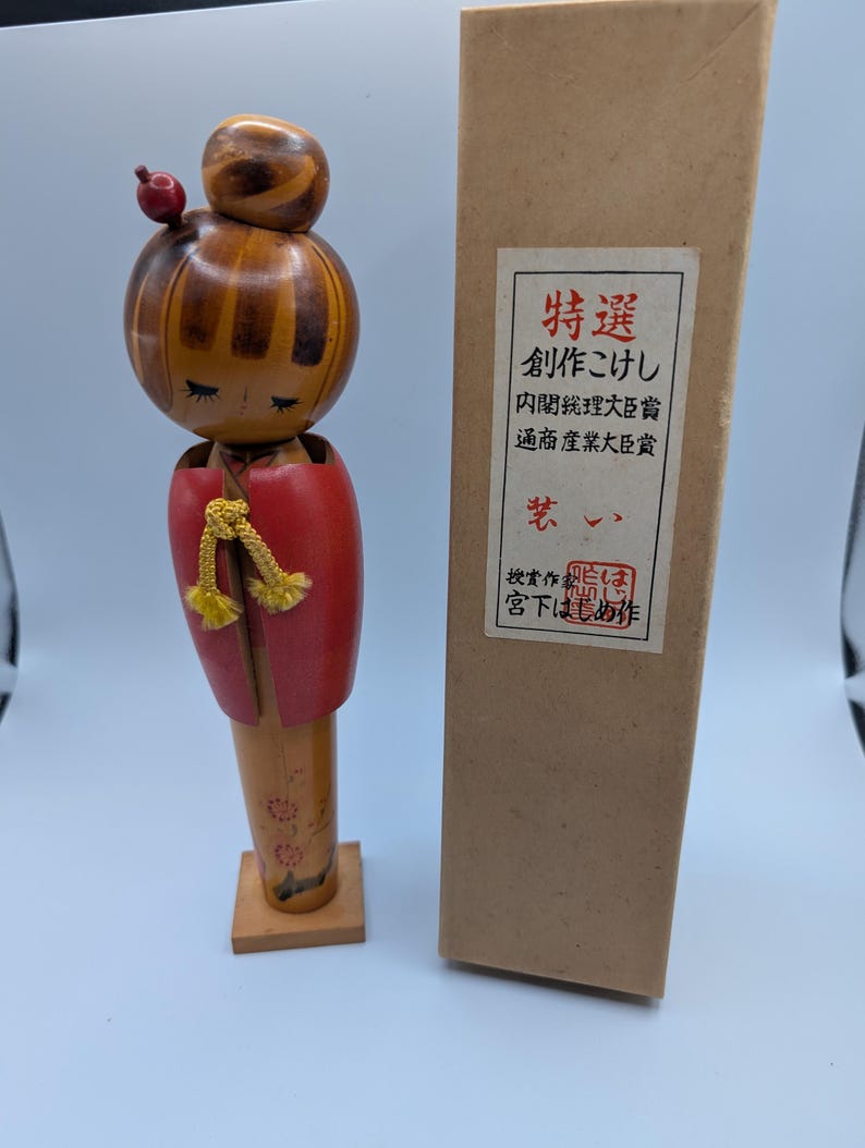 Op de afbeelding: Een houten Japanse Kokeshi-pop met een rode jas en een bruin hoofd. De pop staat naast een bruine doos met Japanse tekst. De pop is ongeveer 15 cm hoog.