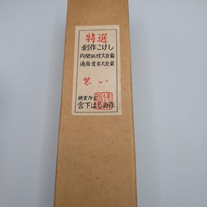 Op de afbeelding: Een lange, rechthoekige kartonnen doos met een label met Japanse tekst en een rode zegel. De doos is lichtbruin van kleur en het label heeft een witte achtergrond met zwarte en rode tekst. De doos lijkt een container te zijn voor een traditioneel Japans ambacht.