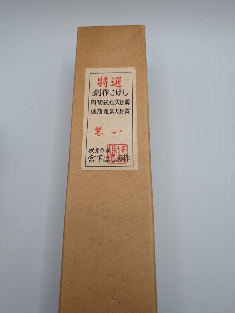 Op de afbeelding: Een lange, rechthoekige kartonnen doos met een label met Japanse tekst en een rode zegel. De doos is lichtbruin van kleur en het label heeft een witte achtergrond met zwarte en rode tekst. De doos lijkt een container te zijn voor een traditioneel Japans ambacht.