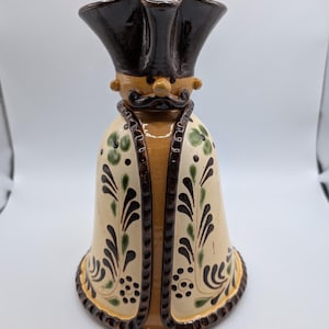 May include: A ceramic pitcher shaped like a figure wearing a dark brown tricorn hat and a mustache. The body is cream-colored with a central tan panel and features hand-painted green and dark brown floral and leaf motifs along the flared base.