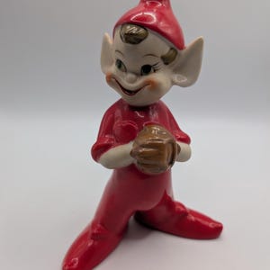 May include: A ceramic elf figurine wearing a bright red pointed hat and a matching red outfit. The elf has large pointed ears, a pale face with painted features, and is holding a small brown baseball glove in both hands with a wide smile.