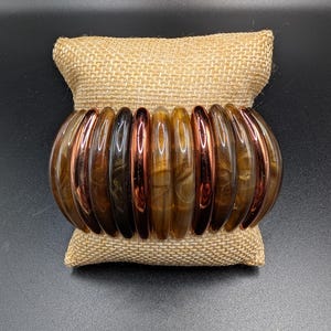 May include: A stretch bracelet composed of alternating cylindrical beads in shades of translucent brown, marbled amber, and metallic copper. The beads are arranged in a continuous loop, creating a rounded, segmented appearance with a polished finish.