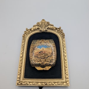 May include: A rectangular metal money clip featuring a black and gold filigree pattern with a central enamel inlay depicting a bridge over water. The money clip is mounted on a black velvet backing inside an ornate, arched gold-tone frame.