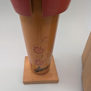 Op de afbeelding: Een houten decoratief item met een rode bovenkant en een lichtbruine body. De body heeft een bloemenmotief in rood. Het item is gemonteerd op een kleine, vierkante houten basis. Een kartonnen doos is gedeeltelijk zichtbaar op de achtergrond.