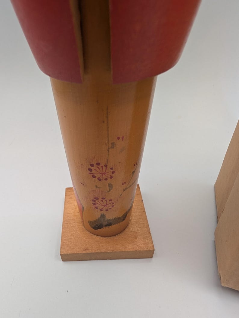 Op de afbeelding: Een houten decoratief item met een rode bovenkant en een lichtbruine body. De body heeft een bloemenmotief in rood. Het item is gemonteerd op een kleine, vierkante houten basis. Een kartonnen doos is gedeeltelijk zichtbaar op de achtergrond.