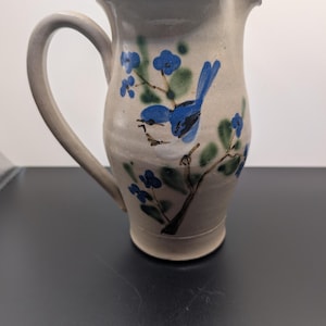 May include: A light beige ceramic pitcher featuring a hand-painted blue bird perched on a brown branch with green leaves and small blue flowers. The pitcher has a curved handle on the left side and a flared spout at the top rim for pouring.