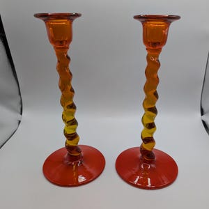 May include: A pair of matching candle holders made of translucent glass. Each holder features a wide, flat circular base in a vibrant orange hue, a tall, slender, spiraled stem that transitions from orange to yellow, and a flared top cup.