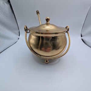 May include: A polished brass pot with a rounded body, three small feet, and a matching lid with a central knob. The pot features a curved swinging handle attached to two side loops and includes a long, thin brass spoon resting inside.