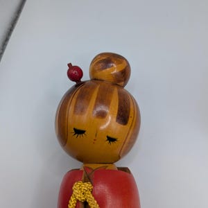 Op de afbeelding: Houten Japanse Kokeshi-pop met een rode body en een gedetailleerde kop. De kop is beschilderd met gesloten ogen en een kleine rode bol zit bovenop de kop. Een goudkleurig koord is om de nek geknoopt.
