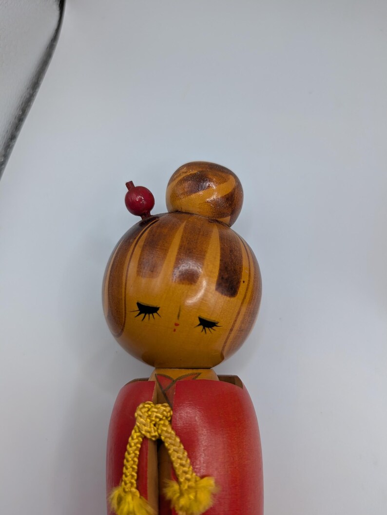 Op de afbeelding: Houten Japanse Kokeshi-pop met een rode body en een gedetailleerde kop. De kop is beschilderd met gesloten ogen en een kleine rode bol zit bovenop de kop. Een goudkleurig koord is om de nek geknoopt.