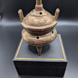 May include: A small, round, metallic bronze-colored incense burner with three legs and two upright handles. The lid and body feature etched patterns and small circular ventilation holes. The burner has a pointed finial on the lid.
