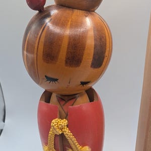 Op de afbeelding: Een vintage houten Kokeshi-pop met een ronde kop en lichaam. De pop heeft een rode top en een gele koord. De kop is beschilderd met bruine en oranje strepen en de pop heeft gesloten ogen. Bovenop het hoofd is een kleine rode bal bevestigd.