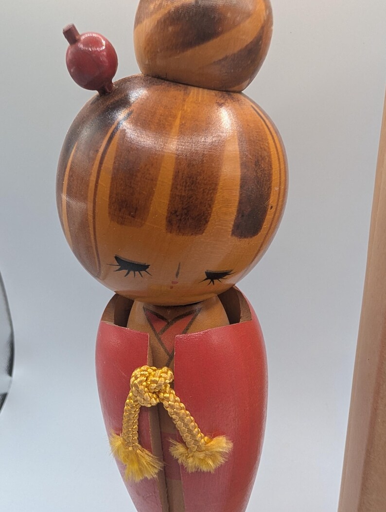 Op de afbeelding: Een vintage houten Kokeshi-pop met een ronde kop en lichaam. De pop heeft een rode top en een gele koord. De kop is beschilderd met bruine en oranje strepen en de pop heeft gesloten ogen. Bovenop het hoofd is een kleine rode bal bevestigd.