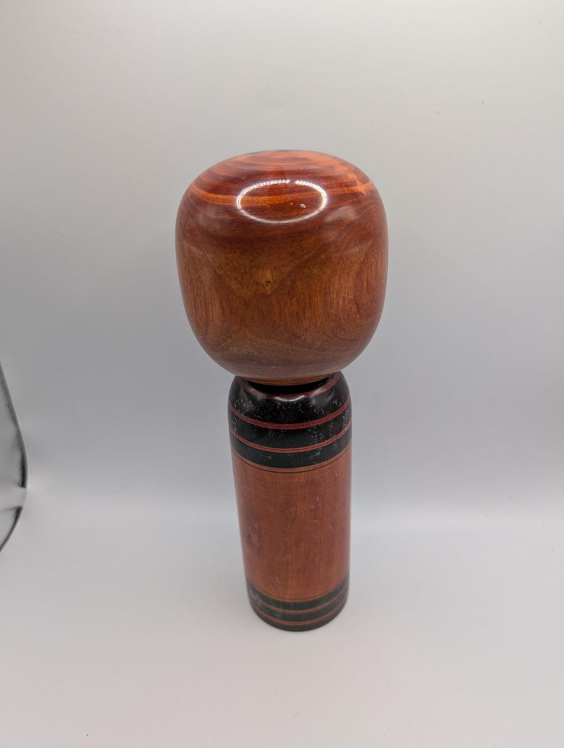 Op de afbeelding: Een gedraaide houten kokeshi-pop met een grote, ronde, gepolijste roodbruine kop en een cilindervormig lichaam. Het lichaam heeft een natuurlijke houtnerfafwerking met horizontale zwarte en donkerrode geschilderde banden.