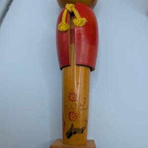 Op de afbeelding: Houten Japanse Kokeshi-pop met een ronde kop en cilindrisch lichaam. De pop heeft een rode jas, gele accenten en bloemmotieven. Hij staat op een kleine houten basis.
