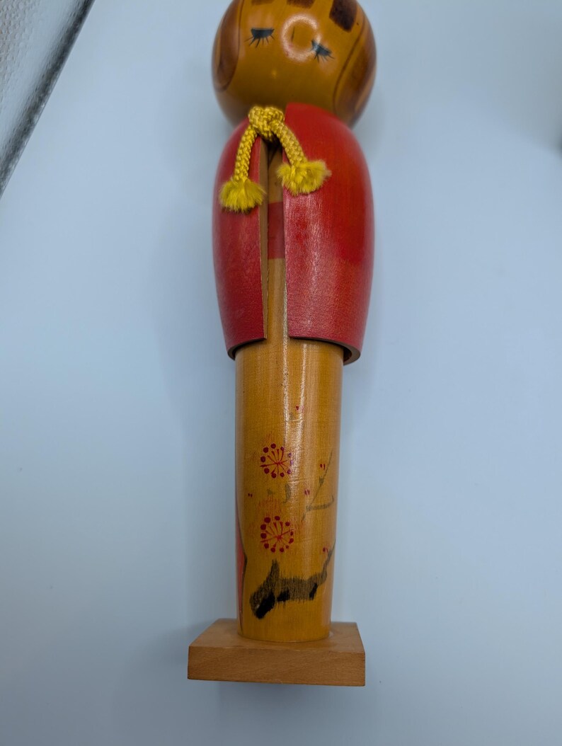 Op de afbeelding: Houten Japanse Kokeshi-pop met een ronde kop en cilindrisch lichaam. De pop heeft een rode jas, gele accenten en bloemmotieven. Hij staat op een kleine houten basis.