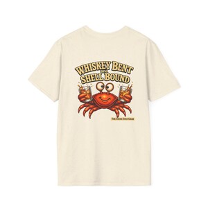 May include: A cream-colored t-shirt featuring a graphic of a smiling orange crab holding two glasses of whiskey. The text above the crab reads "WHISKEY BENT AND SHELL BOUND" in a bold font, and the text below reads "THE CROSS EYED CRAB".