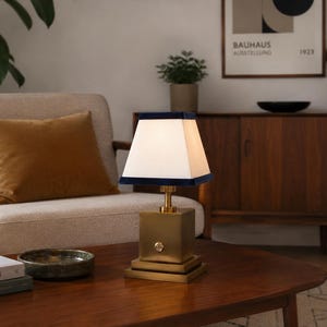 May include: A table lamp with a square brass base and a white tapered lampshade featuring a dark blue velvet trim at the top and bottom edges. The lamp has a metallic finish and a small circular switch on the front of the square base.