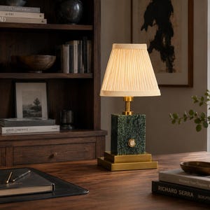 May include: A table lamp featuring a rectangular green marble base with a brass finish square pedestal and a matching brass neck. The lamp is topped with a cream-colored, pleated, conical fabric shade that emits a warm, soft glow.