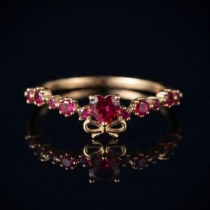 May include: A gold ring featuring a central heart-shaped red gemstone flanked by smaller round red gemstones along the band. A small gold bow detail sits directly beneath the heart-shaped stone, adding a decorative element to the metal band.