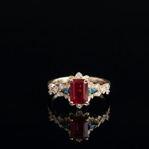 Puede incluir: Un anillo de oro con una piedra roja central de corte esmeralda, flanqueada por dos piedras de color verde azulado en corte de pera. La banda está decorada con pequeñas piedras claras de corte redondo en un diseño de metal texturizado y orgánico.