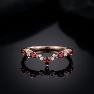 May include: Rose gold ring with a curved design. The ring features a series of small, round white gemstones and larger, teardrop-shaped red gemstones. The ring is set against a dark background, with its reflection visible below.