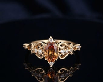 Puede incluir: Un anillo de oro con una piedra preciosa naranja de corte ovalado en el centro, rodeada de pequeñas piedras claras. La banda presenta detalles metálicos intrincados en forma de volutas a ambos lados del engaste principal.