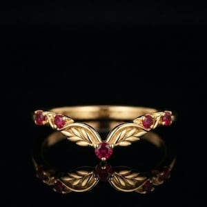 Può includere: Un anello in oro a forma di V con cinque rubini rossi rotondi incastonati lungo la fascia. La banda metallica è scolpita con un motivo a foglie dettagliato che si estende dal rubino centrale verso le estremità, creando un design simmetrico.