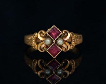 May include: A gold ring featuring two square-cut deep red gemstones arranged vertically, flanked by two small round white pearls. The metal band displays intricate scrollwork and decorative detailing on both sides of the central setting.