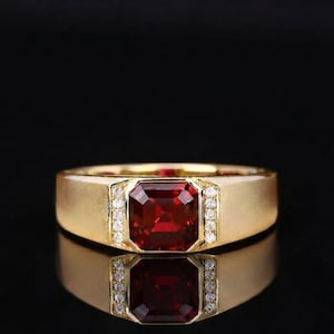May include: A gold ring featuring a central emerald-cut deep red gemstone. The stone is flanked on both sides by a vertical row of three small, clear, round-cut accent stones set into the band. The band has a smooth, polished finish.