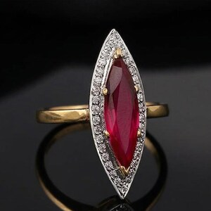 Op de afbeelding: Een gouden ring met een marquise-vormige, dieprode edelsteen, omringd door een halo van kleine diamanten. De ring heeft een gepolijste gouden band, die contrasteert met de rode edelsteen en de diamanten.