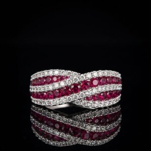 May include: A crossover ring featuring alternating rows of round-cut red rubies and clear white diamonds set in a polished metal band. The design consists of two intertwined bands that create a continuous wave pattern across the finger.