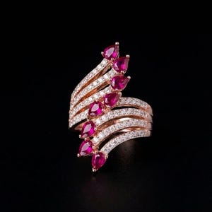 May include: A rose gold ring featuring a bypass design with multiple bands encrusted with small white stones. The ring is accented by a series of seven pear-shaped, deep pinkish-red gemstones arranged in a diagonal line across the centre.