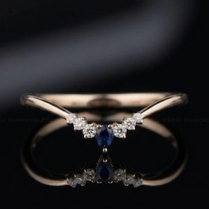 May include: A rose gold chevron ring featuring a central pear-shaped blue sapphire flanked by three small round white diamonds on each side, creating a symmetrical V-shaped design that rests on a reflective surface against a dark background.