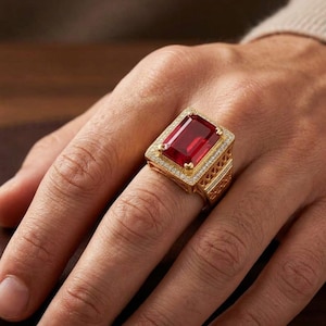 May include: A gold ring featuring a large, rectangular, faceted red gemstone set in the center. The stone is surrounded by a halo of small white stones, with additional white stones embedded along the sides of the gold band.