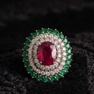 May include: A ring featuring a central oval-cut red ruby surrounded by two concentric halos of small round white diamonds and an outer border of pear-cut green emeralds set in a metallic band with a multi-layered floral design.
