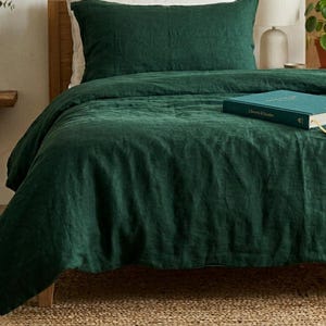 Dark emerald green linen bedding set on a wooden bed in a minimalist bedroom with natural wood furniture, soft neutral decor, and warm natural light.
